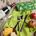 Wine bottle, glass, cheese, bread, apples, and cutlery on a green cloth with an avocado-patterned bag.