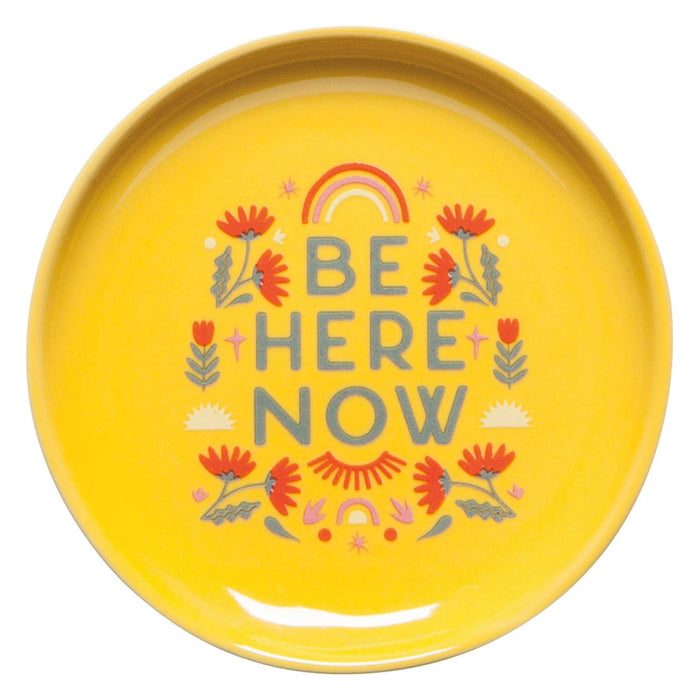 Yellow plate with 'Be Here Now' text and floral design on a white background