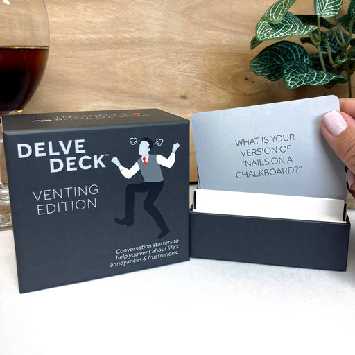 Delve Deck venting edition card game with a card held by a hand, placed on a wooden surface with a plant in the background.