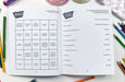 Open book with a bingo-style game on a table with colored pencils